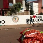 Long line of voters waiting outside a polling station in Bissau on election day 2025, with security personnel visible and national flags in the background