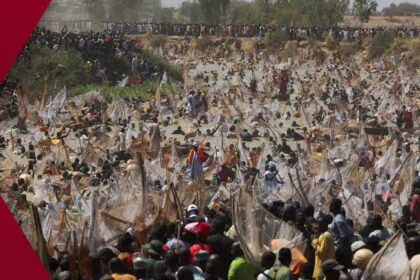 Dozens of Nigerian men in colorful shorts sprint and splash into the muddy Matawalle River at the 2026 Argungu Fishing Festival, holding large handmade nets, while spectators watch from the riverbank under a hazy sky.